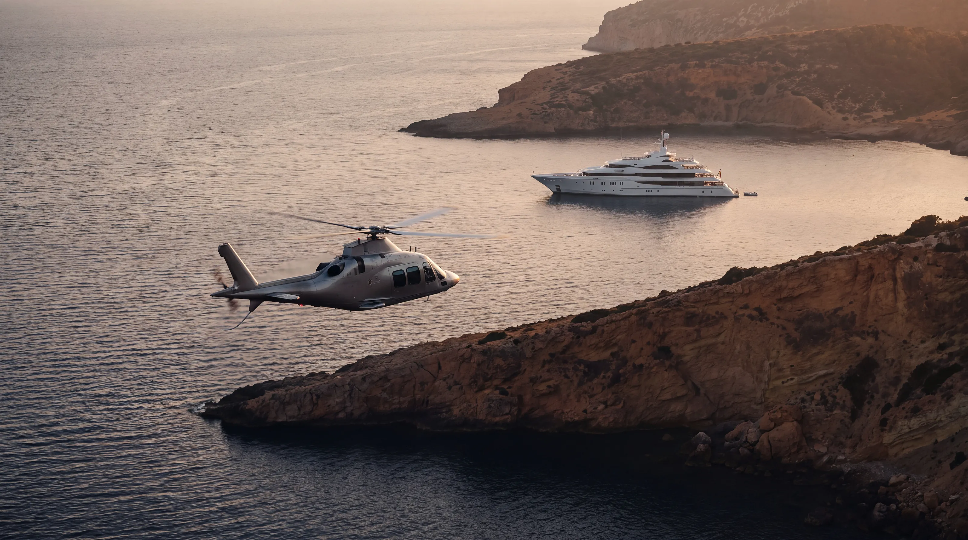 A silver helicopter flies along a coastline toward a superyacht at anchor at dusk